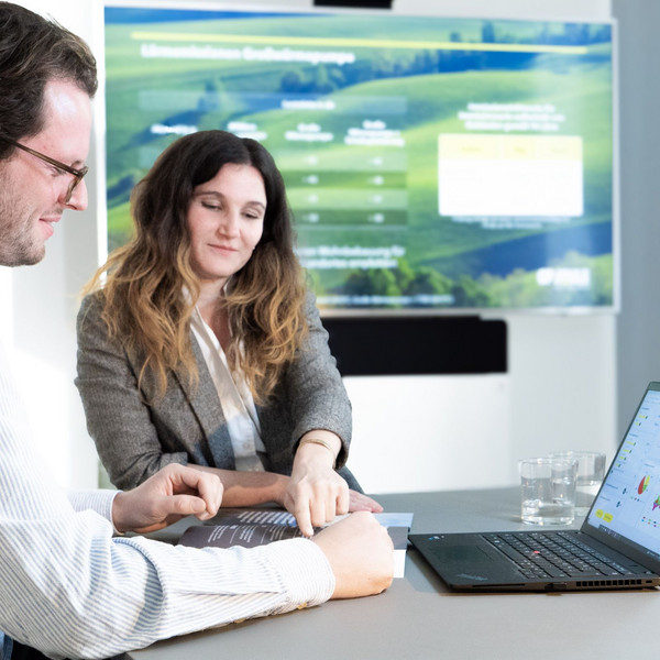 Drei Personen sitzen im Besprechungsraum, betrachten Unterlagen und Auswertungen auf einem Laptop vor einem großen Präsentationsbildschirm.