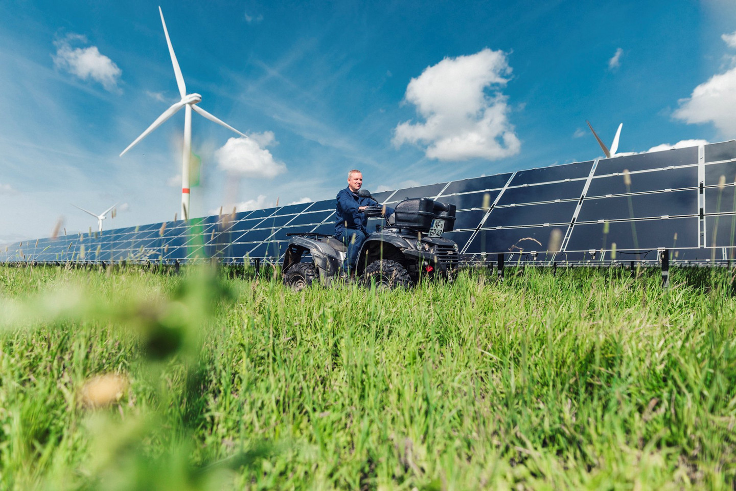 Mitarbeiter fährt auf Quad durch Solarfeld, im Hintergrund Windräder und blauer Himmel mit Wolken.
