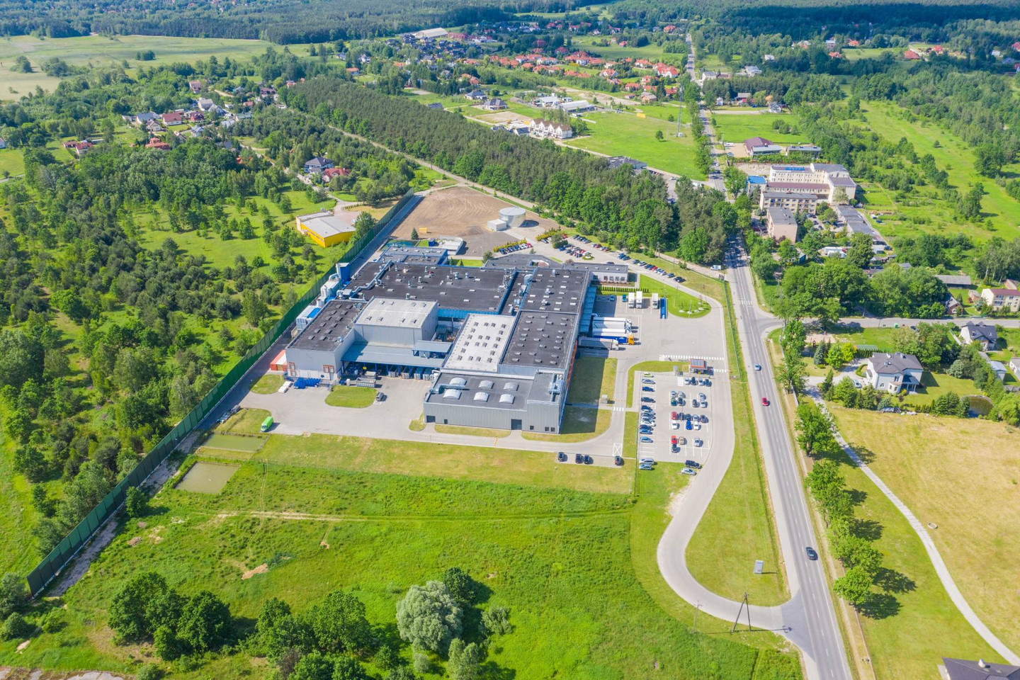 Luftaufnahme eines Industriegebäudes mit Parkplatz, umgeben von Straßen, Wiesen, Wald und verstreuten Wohnhäusern.